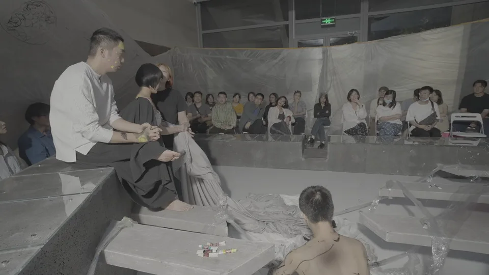 Performers sit on stepped gray platforms while spectators watch from the same visible field in Human Surrender.