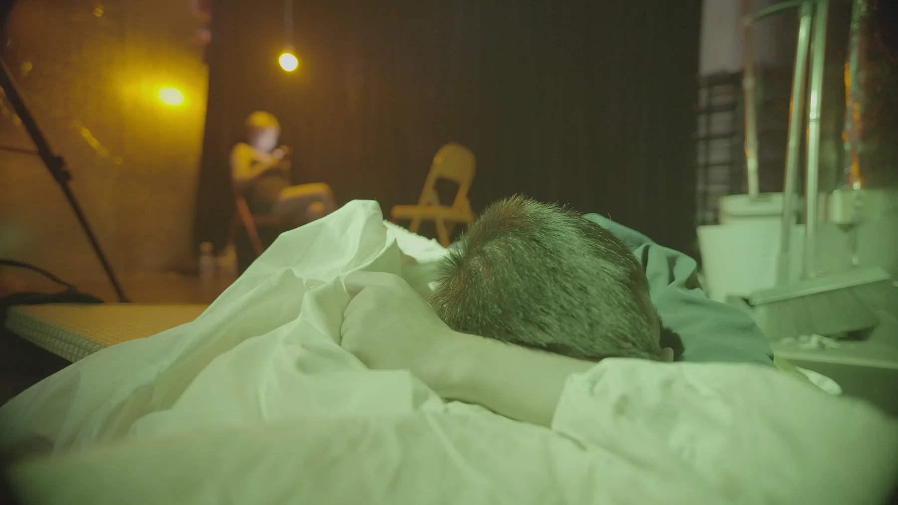 A person lies face down among white bedding while a seated figure appears out of focus beneath warm lights in the background.